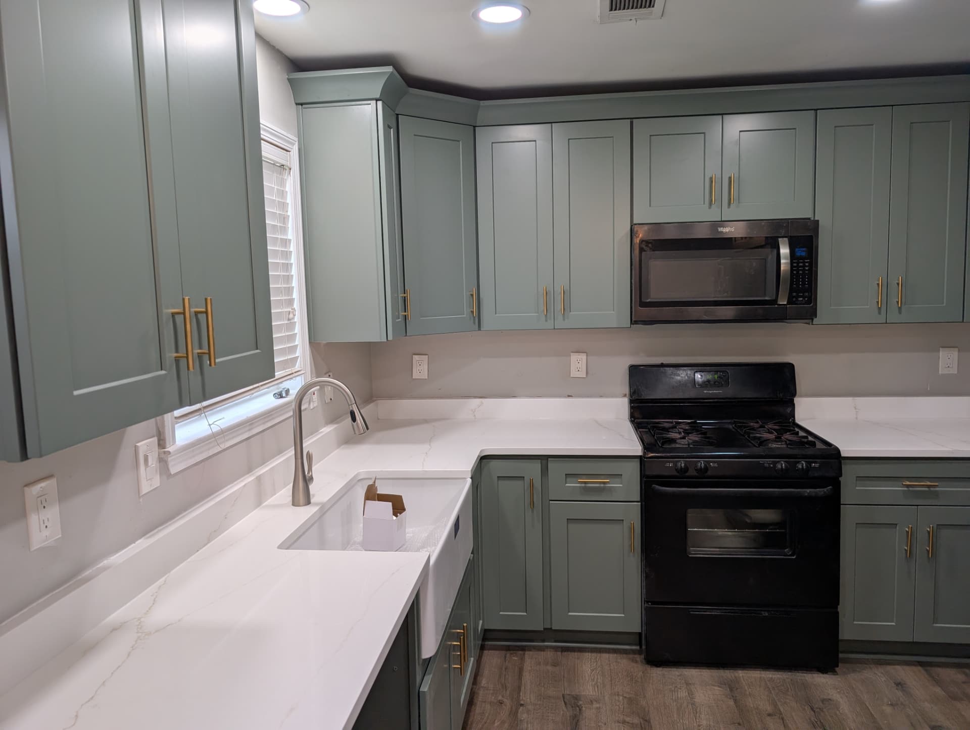 Muted green-gray shaker cabinets with gold-tone pulls, white lightly veined countertops, an apron-front sink beneath a window, and black appliances.